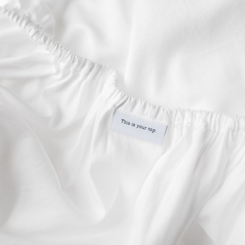 Close up of Tuck's crisp white bedsheet. A tag reads, "This is your top."