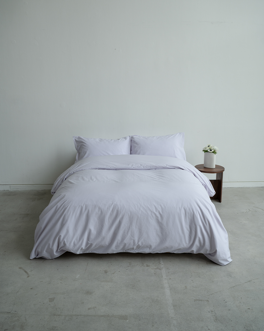 Lavender bedding set on a bed against a plain wall with a small table and plant in the background.
