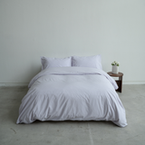Lavender bedding set on a bed against a plain wall with a small table and plant in the background.